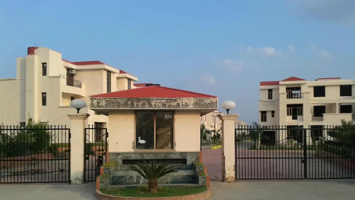 Samiah Media Village — A security gate and booth at the entrance of a residential complex with cream-colored buildings and red roofs under a blue sky.